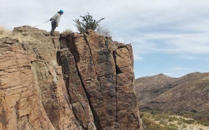 a rock climber who is secured by ropes stands on a cliff and leans over the edge to get a better look